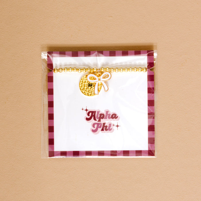 The Darling Effect’s Alpha Phi Bow Gold Charm Bracelet features a gold beaded design with a round textured charm and bow, packaged on an Alpha Phi card with pink letters and a checkered border—perfect for showing chapter pride.