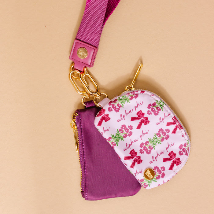 The Darling Effect's Alpha Phi Dynamic Duo Pouch Wristlet features two zippered pouches—one solid purple, one with Alpha Phi, pink flowers, and maroon umbrellas—plus a purple lanyard with gold hardware for hands-free convenience.