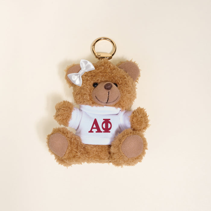 The Darling Effect’s Alpha Phi Teddy Bear Bag Charm is a small plush bear with a white bow and shirt featuring red ΑΦ letters, perfect as a cute sorority gift or accessory for your bag.