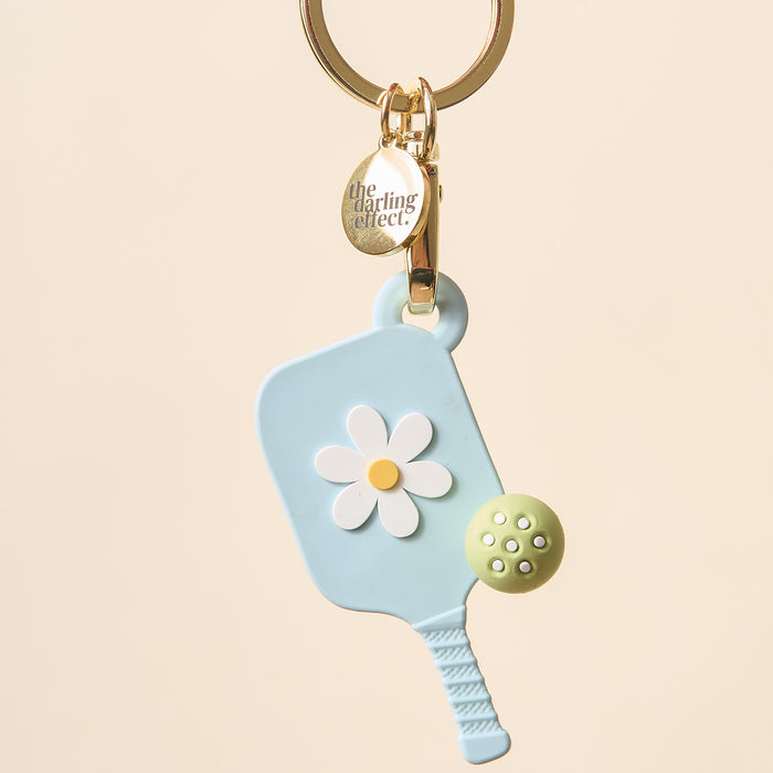 The Darling Effect's Aqua Pickleball Paddle Keychain features a pastel blue paddle with a white and yellow daisy, a green ball, and a gold charm—making it the perfect unique gift for pickleball lovers.