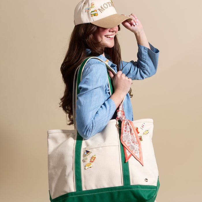 A woman in a light blue shirt and beige cap smiles while holding her hat's brim. Her large cream tote bag with green trim is decorated with The Darling Effect's Berrylicious The Pin Pack and has a patterned scarf tied to the handle.