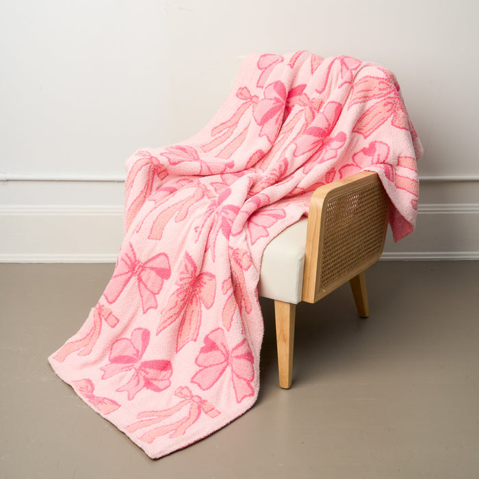 The-darling-effect's Blushing Bows Pink Cozy Dreams Luxe Blanket, made from ultra-soft polyester microfiber with pink bow patterns, is draped over a light wood and cane chair with a white cushion against a beige floor and white wall.