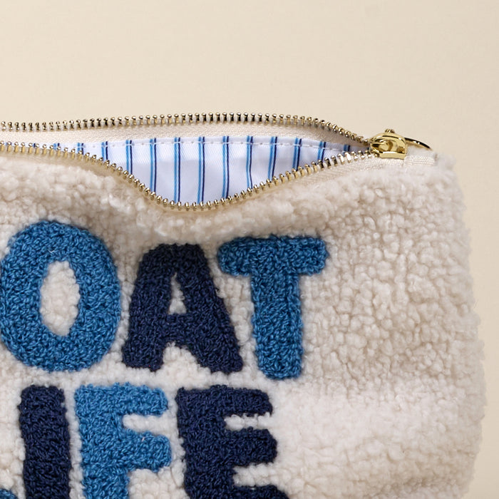 Close-up of the-darling-effect’s Boat Life Teddy Pouch in cream sherpa with blue “BOAT LIFE” embroidery. A partially open zipper reveals its cozy blue and white striped lining.