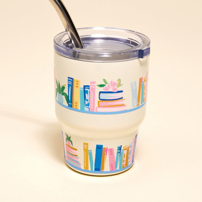 The Darling Effect’s Book Shelfie 2 oz Tiny Tumbler features colorful bookshelf illustrations, a cream base, clear lid, and metal straw—perfect as a mini tumbler or for your next espresso shot.