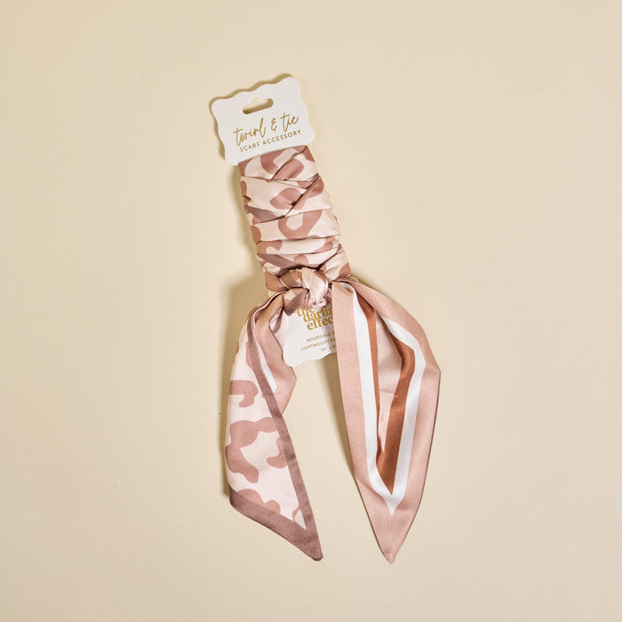 The Darling Effect’s Born Wild Twirl & Tie Scarf Accessory is a soft pink and beige animal print scrunchie with an attached ribbon, shown on cream with a “twirl & tie” label—perfect for unlimited styling.