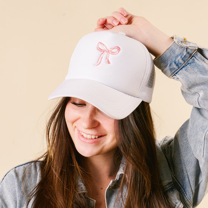 A woman in a denim jacket wears the Bow Pink Trucker Hat by the-darling-effect, smiling as she holds its brim against a beige background—showcasing how this trendy hat can elevate any look.