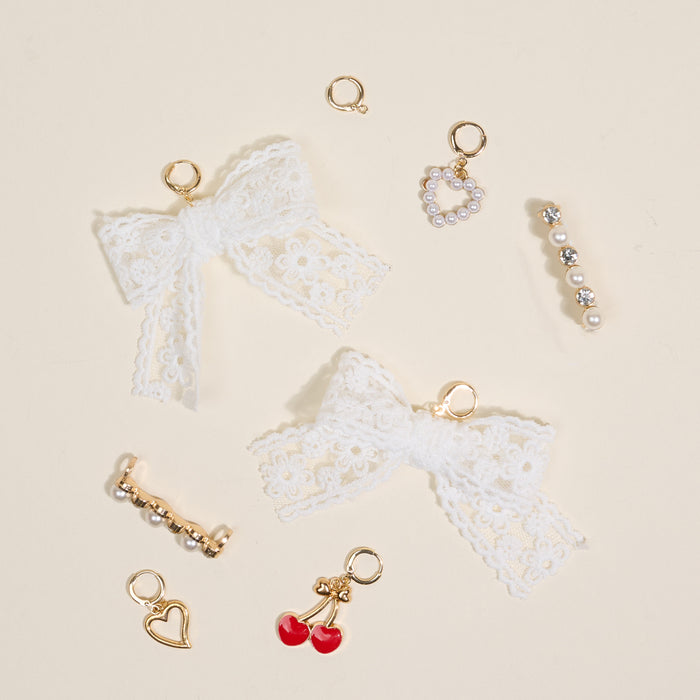 A flat lay of the-darling-effect’s Cherry Twist Dazzle Shoe Charm Pack—featuring white lace bows, a gold and pearl heart, cherry charm, and gold rings—makes unique sneaker or footwear accessories on a light beige background.