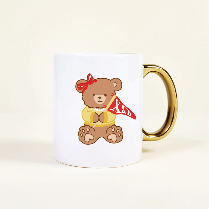 The Darling Effect's Chi Omega 11 oz Cozy Cute Ceramic Mug features a brown teddy bear in a red bow and yellow shirt, holding a red pennant with white Greek letters, and has a charming gold handle.