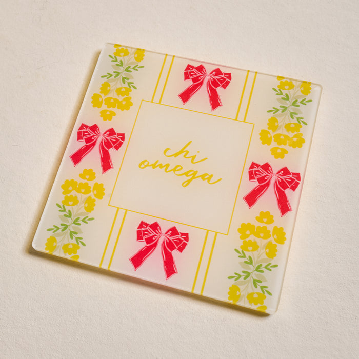 The Darling Effect’s Chi Omega Acrylic Coaster features a white square design with yellow floral patterns, red bows at the edges, and "chi omega" in yellow cursive—ideal as a sorority coaster or thoughtful gift.