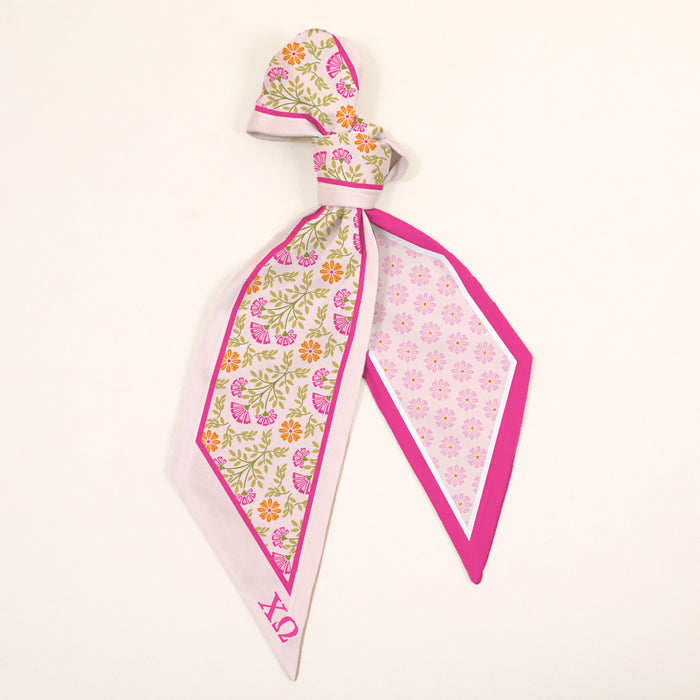 The Darling Effect's Chi Omega Twirl & Tie Scarf Accessory is a silk scarf with a pink, yellow, and green floral pattern on cream, pink border, and OX in pink. Perfect for versatile styling and sorority spirit.