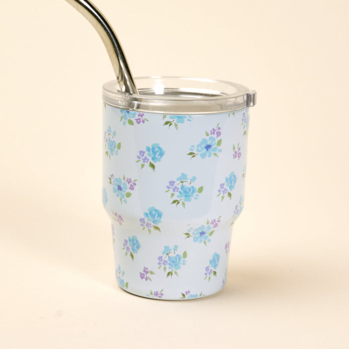 The Dainty Floral Blue 2 oz Tiny Tumbler by the-darling-effect features a light blue design with blue and purple flowers, a clear lid, and a metal straw, photographed on a plain beige background.