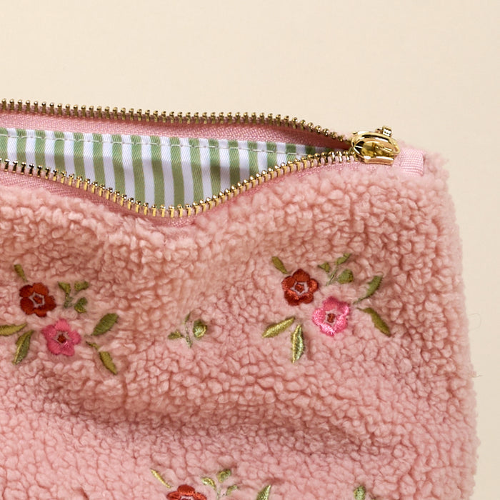 A close-up of the-darling-effect's Dainty Floral Pink Teddy Pouch, featuring pink sherpa with embroidered red and pink flowers, a gold zipper, and a green and white striped fabric lining inside.