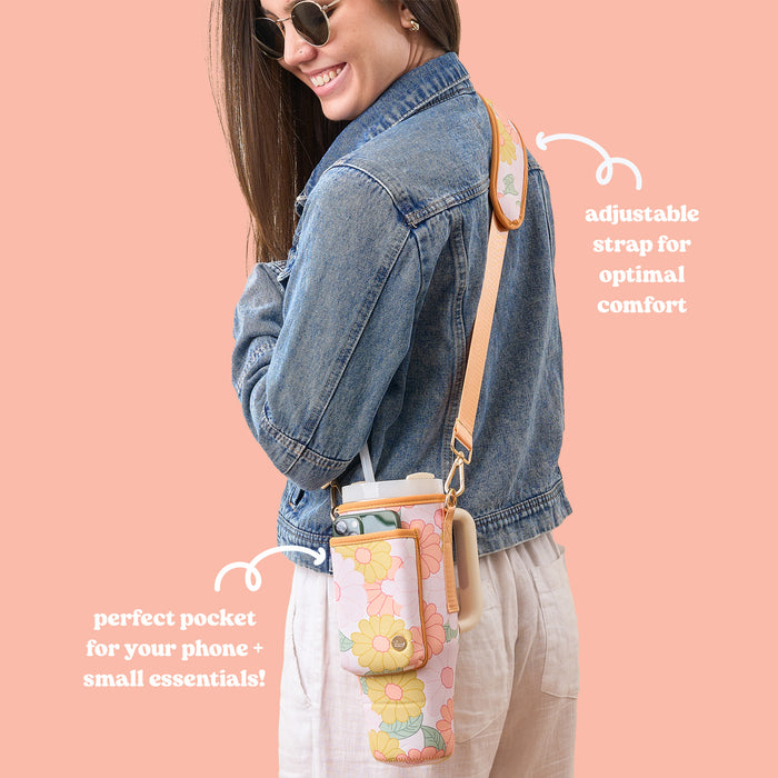 A smiling woman in sunglasses, a denim jacket, and light pants carries The Darling Effect’s Daisy Craze Peach Tumbler Sling—a floral neoprene sling bag with an adjustable strap, phone pocket, and versatile features.
