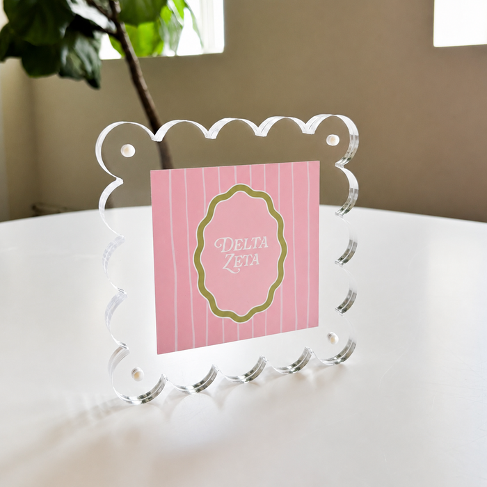 The Darling Effect’s Delta Zeta Square Scalloped Acrylic Frame displays a pink Delta Zeta card on a white table with a plant and window—ideal for fans of modern decor.