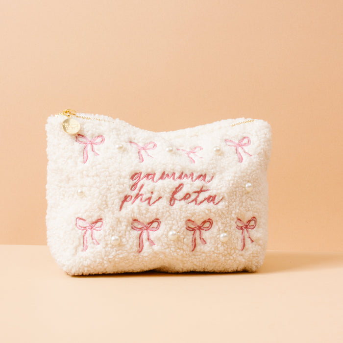 The Darling Effect’s Gamma Phi Beta Bows & Pearls Teddy Pouch is a cream sherpa zippered pouch with pink bows, pearls, and "Gamma Phi Beta" embroidered on the front—perfectly giftable for sorority sisters.