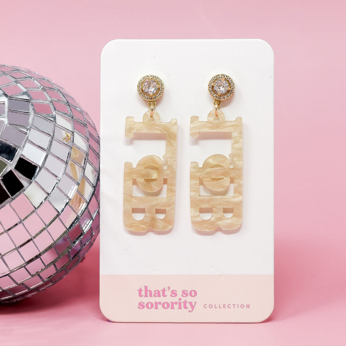 A pair of Gamma Phi Beta Letter Gold Acrylic Earrings by The Darling Effect rests on a card that reads "thats so sorority COLLECTION," set against a pink background with a silver disco ball—ideal for sisterhood events or bid-day baskets.