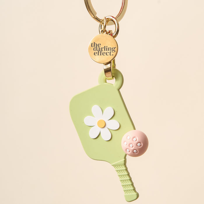 The Darling Effect’s Green Pickleball Paddle Keychain features a light green paddle with a white daisy, a pink ball, and a gold keyring with an engraved charm—making it the perfect pickleball gift.