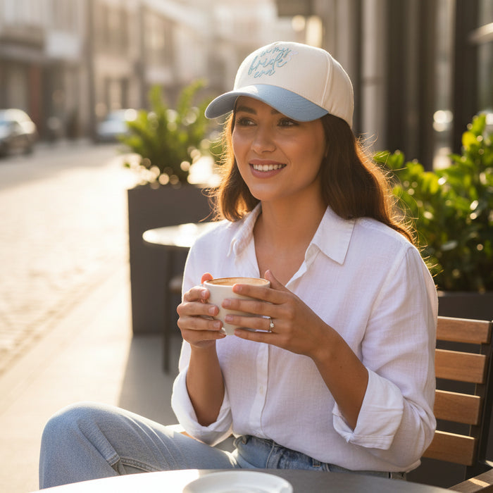 A woman in a white shirt and The Darling Effect's In My Bride Era Classic Canvas Hat sits at an outdoor café table, smiling with coffee in hand and city views behind her, showcasing a relaxed, everyday look.