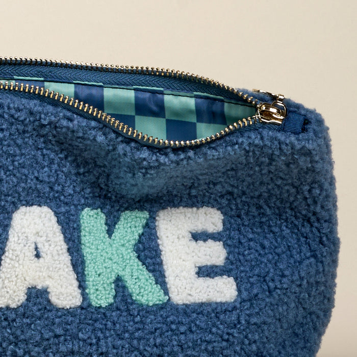 The Lake Teddy Pouch by the-darling-effect is a blue sherpa pouch with a zippered top, partially open to show its blue and green checkered lining and large white and light green "AKE" letters—a cozy pick for teddy pouch fans.