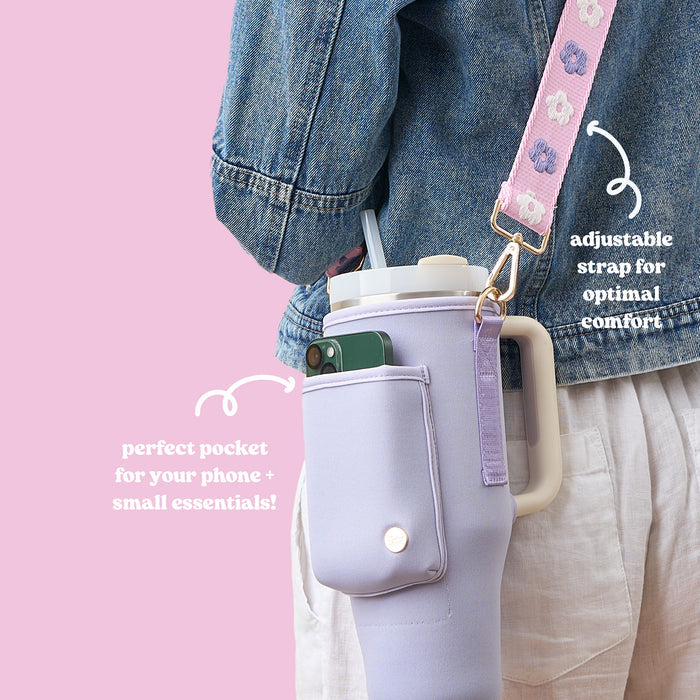 A person carries The Darling Effect’s Periwinkle Tumbler Sling—a neoprene bag with a lavender sleeve, phone pocket, and pink floral adjustable strap—pairing it with a denim jacket and linen pants for versatile style and function.