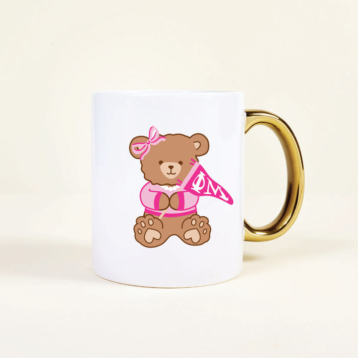 The Darling Effect’s Phi Mu 11 oz Cozy Cute Ceramic Mug is a white ceramic mug with a gold handle, featuring a brown bear in a pink bow and cardigan holding a flag with ΦM—perfect for showing sorority spirit.
