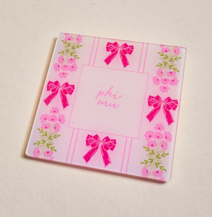 The Darling Effect's Phi Mu Acrylic Coaster is a square pink coaster featuring "phi mu" in the center with bows and floral edges—ideal for chapter pride or as a cute Big/Little gift, beautifully displayed on a light surface.