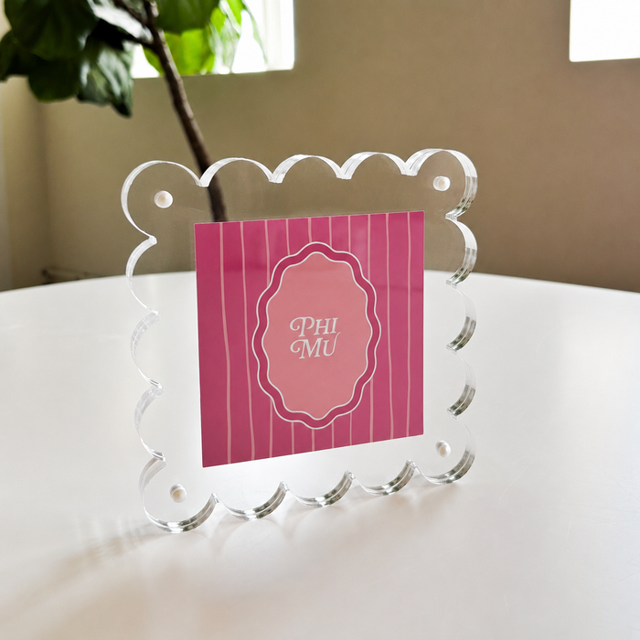 The Darling Effect's Phi Mu Square Scalloped Acrylic Frame displays a pink PHI MU card in white script on a table with a leafy plant and window, adding charm to your modern picture frames collection.