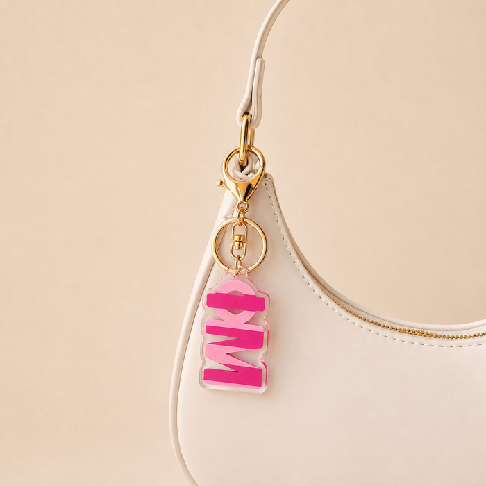 A close-up of a white handbag with a gold clasp holding The Darling Effect’s Phi Mu Stripes Acrylic Keychain—shaped like the word MOM in block letters. The background is a soft beige, making it a perfect sorority gift.