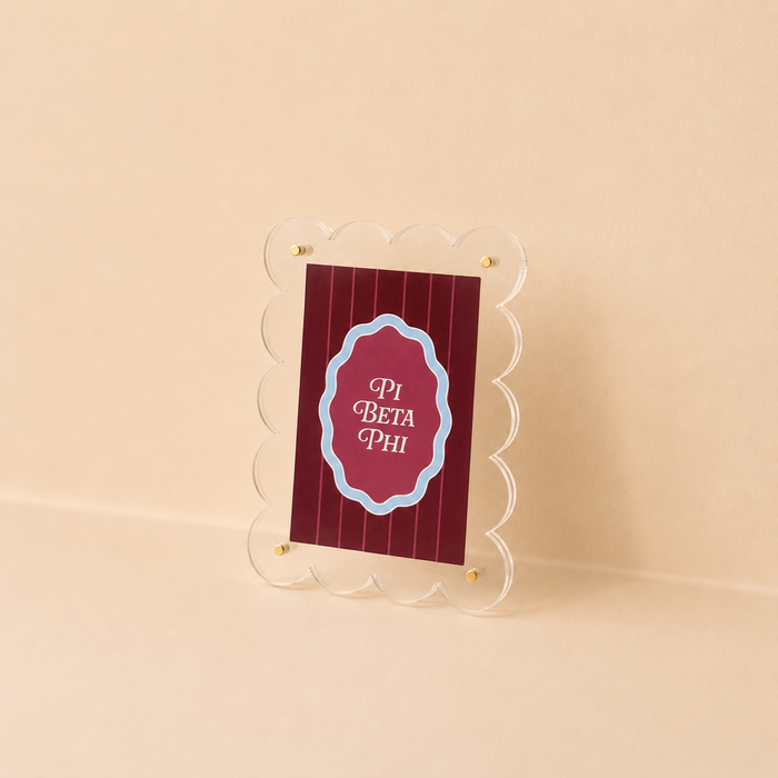 The Darling Effect Pi Beta Phi Mini Acrylic Picture Frame Magnet displays a maroon card with vertical stripes and "Pi Beta Phi" in white text bordered in blue. The small, scalloped, clear frame stands on a light beige surface and background.