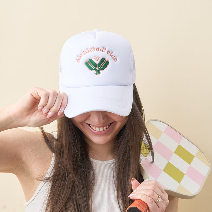 A smiling woman wears the-darling-effect's Pickleball Club Trucker Hat with "pickleball club" and crossed paddles embroidered on front. She holds a pink and green checkered pickleball paddle.