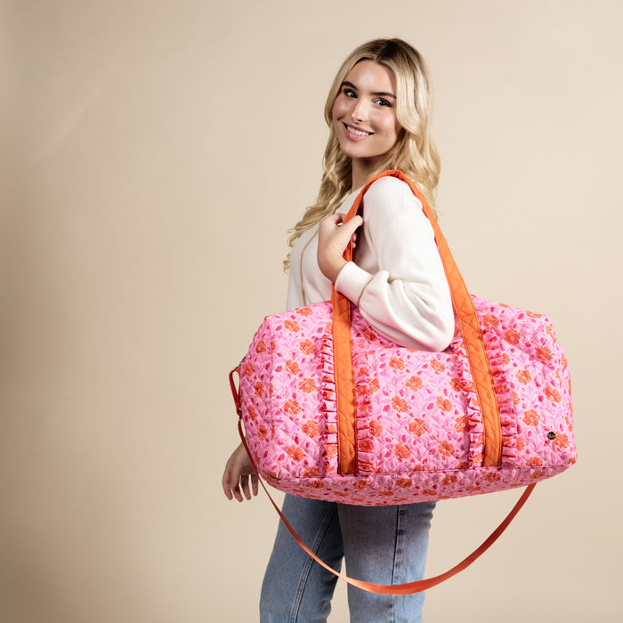 A blonde woman in a white sweater and jeans carries The Darling Effect’s Poppin’ Petals Hot Pink On-the-Go Quilted Weekender Bag, featuring an orange floral pattern and orange ruffled straps.