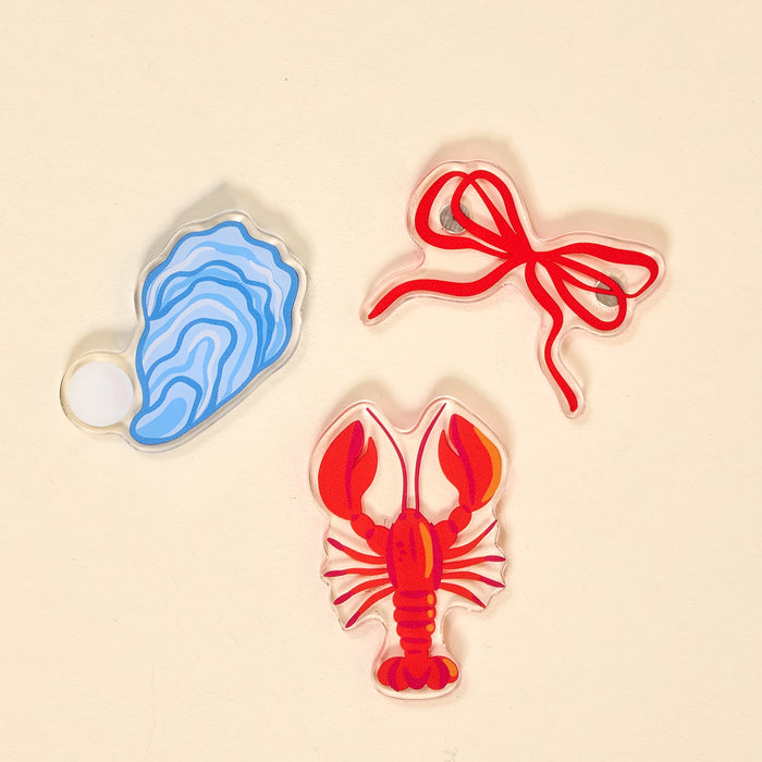 The Darling Effect’s Seaside Haven Little Magnet Set includes a blue oyster with pearl, red lobster, and red tied ribbon acrylic pins on beige—charming accents that double as unique fridge magnets.