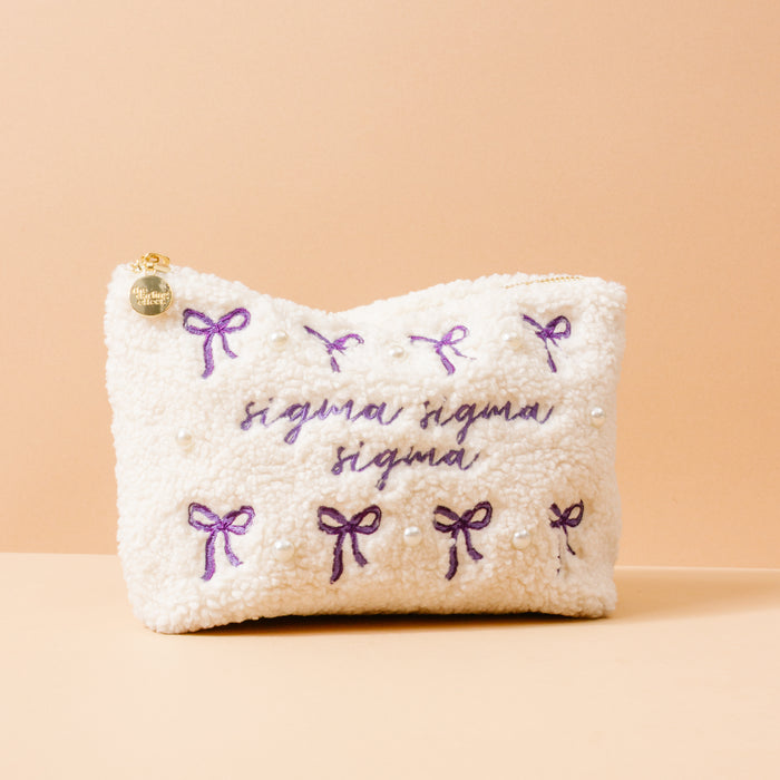 The Darling Effect's Sigma Sigma Sigma Bows & Pearls Teddy Pouch is a fluffy white sherpa pouch with purple embroidered bows, pearls, Tri Sigma text, and a gold zipper—perfect as a sorority gift.