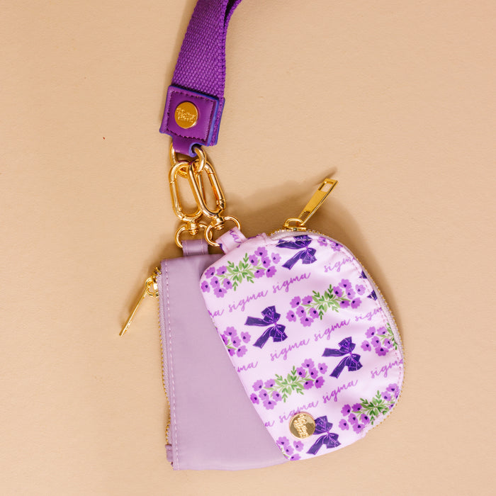 The Darling Effect’s Sigma Sigma Sigma Dynamic Duo Pouch Wristlet is a small pastel purple purse with gold zipper and hardware, a matching strap, and a bows, flowers, and “sigma” cursive pattern on a light background for hands-free style.
