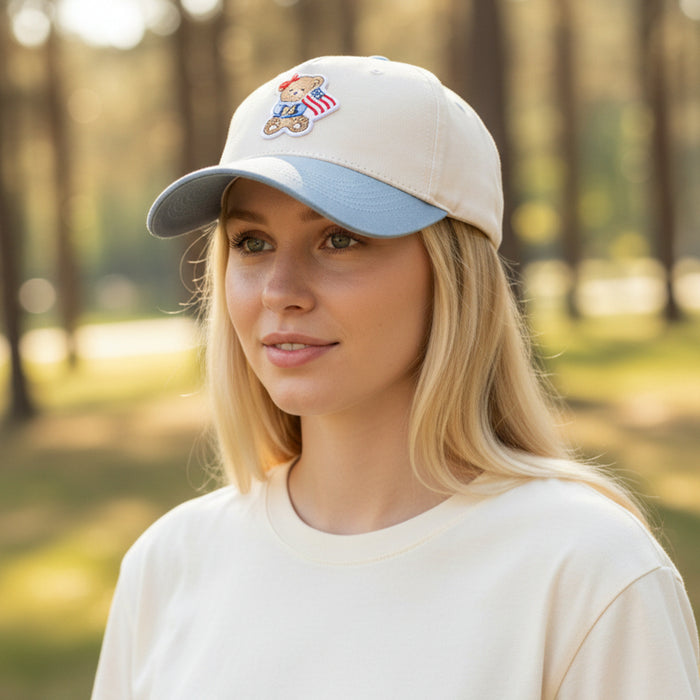 A young woman with long blonde hair stands outdoors in a relaxed light T-shirt and The Darling Effect's Star Spangled Bear Classic Canvas Hat, featuring a bear and American flag patch, with blurred trees behind her.