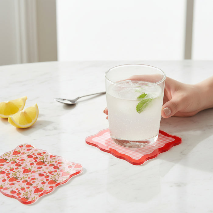 A hand holds a glass of sparkling water with ice and mint on The Darling Effect's Strawberry Fields Table Talk Acrylic Coaster, surrounded by lemon wedges, a spoon, and stylish décor atop a white marble surface.