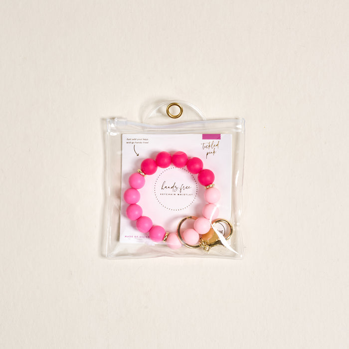 The Darling Effect Tickled Pink Hands-Free Keychain Wristlet, a beaded pink and white silicone key holder with gold accents, is shown in a clear pouch with a gold grommet. The pouch includes an insert reading "lady flow" and "Twisted pink.
