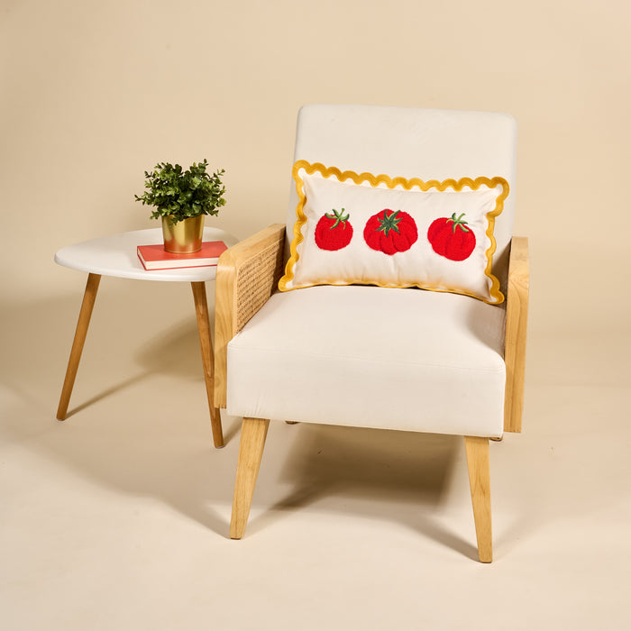 A white and wooden armchair with The Darling Effect’s Tomatoes Scalloped Rectangle Throw Pillow sits beside a small white side table holding a potted plant and two books, all set against a beige background.