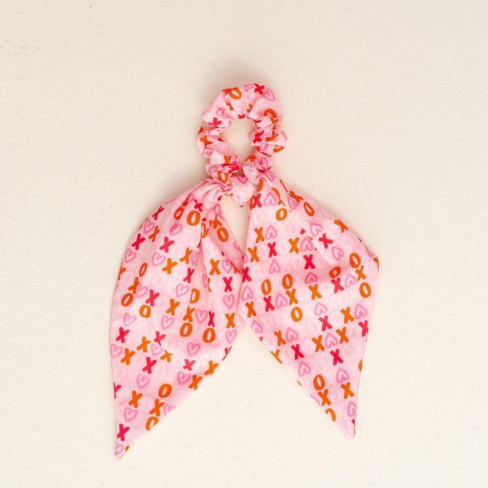 The XOXO Hair Scarf by the-darling-effect is a pink scrunchie with two scarf-like tails, displaying an XOXO pattern in pink, red, and orange on a plain off-white background.