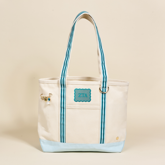 The Darling Effect Zeta Tau Alpha Canvas Tote Bag features cream canvas, light blue accents, blue and white striped handles, a turquoise ZTA patch, and a decorative pin—perfect as a stylish sorority tote or thoughtful gift. Beige background.