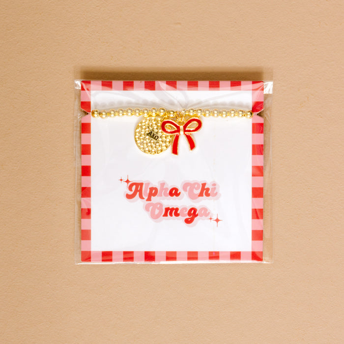 The Darling Effect's Alpha Chi Omega Bow Gold Charm Bracelet features gold beads, a round charm, and red bow on a display card with pink Alpha Chi Omega script, red gingham border, and tan background—perfect for showing your sorority pride.