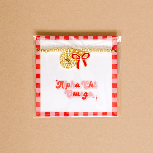 The Darling Effect's Alpha Chi Omega Bow Gold Charm Bracelet features gold beads, a round charm, and red bow on a display card with pink Alpha Chi Omega script, red gingham border, and tan background—perfect for showing your sorority pride.