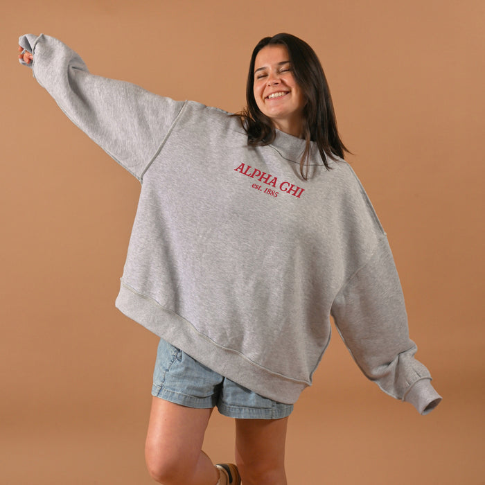 A smiling woman with long brown hair wears The Darling Effect’s Alpha Chi Omega Chapter Mockneck Sweatshirt (L/XL, gray), styled oversized with denim shorts, standing arms outstretched against a plain tan background.