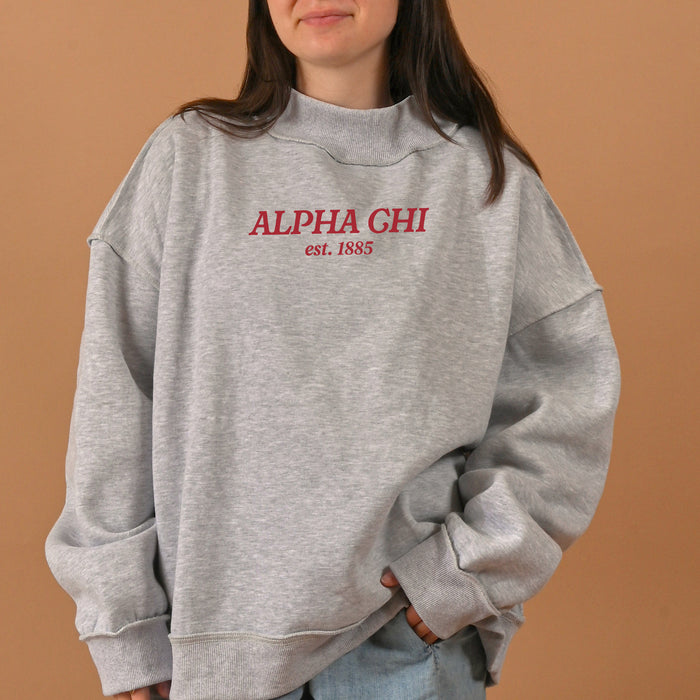 A person stands against a plain tan background wearing The Darling Effect's Alpha Chi Omega Chapter Mockneck Sweatshirt in size L/XL, featuring "ALPHA CHI est. 1885" printed in red text.