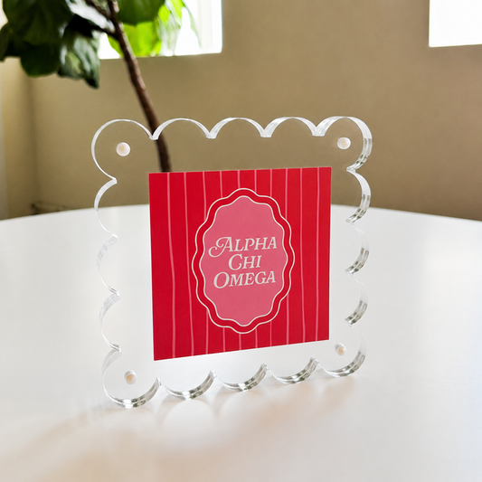 The Darling Effect's Alpha Chi Omega Square Scalloped Acrylic Frame displays a red Alpha Chi Omega card on a white table, with green leaves and windows softly blurred in the background.
