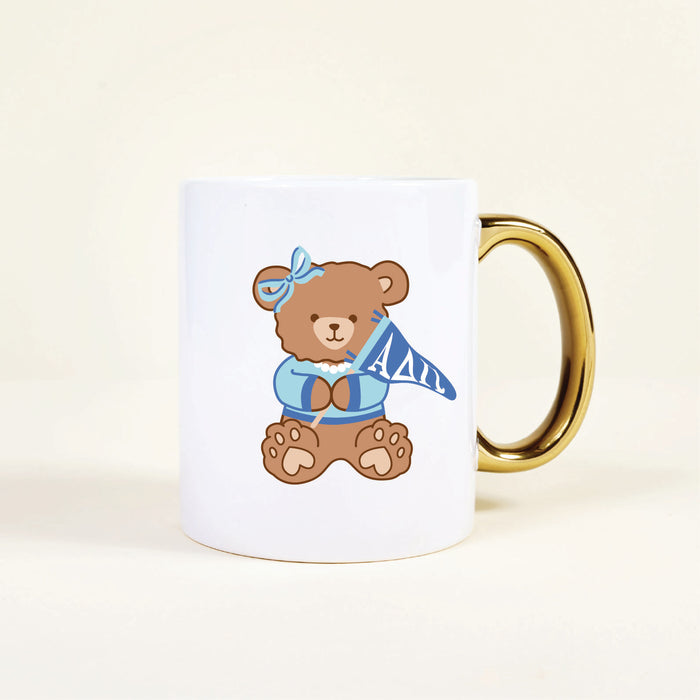 The Darling Effect’s Alpha Delta Pi 11 oz Cozy Cute Ceramic Mug features a brown teddy bear in a blue bow and sweater holding an ΑΔΠ pennant, plus a shiny gold handle for extra charm.