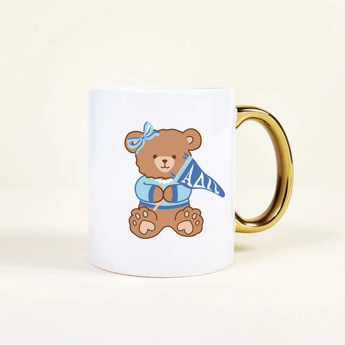 The Darling Effect’s Alpha Delta Pi 11 oz Cozy Cute Ceramic Mug features a brown teddy bear in a blue bow and sweater holding an ΑΔΠ pennant, plus a shiny gold handle for extra charm.