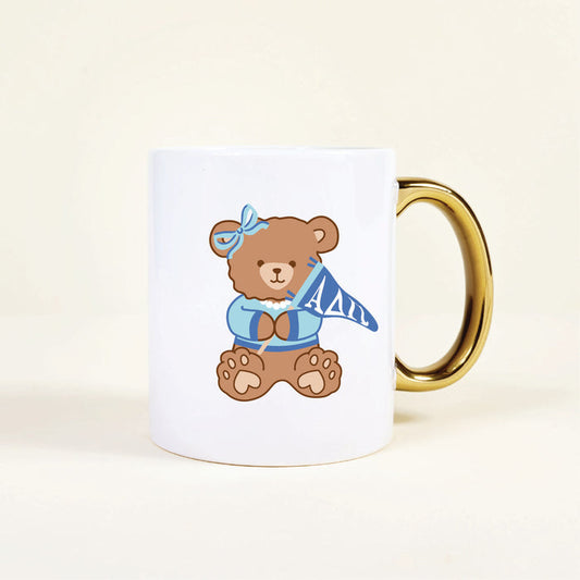 The Darling Effect’s Alpha Delta Pi 11 oz Cozy Cute Ceramic Mug features a brown teddy bear in a blue bow and sweater holding an ΑΔΠ pennant, plus a shiny gold handle for extra charm.