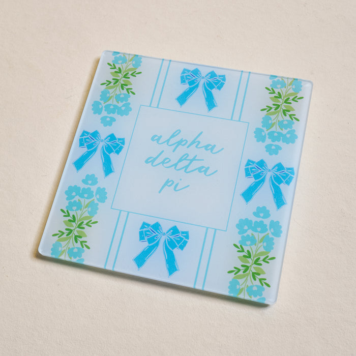 The Darling Effect’s Alpha Delta Pi Acrylic Coaster features "alpha delta pi" in light blue with blue bows and green-blue florals on white—a stylish gift for Bigs or Littles!.