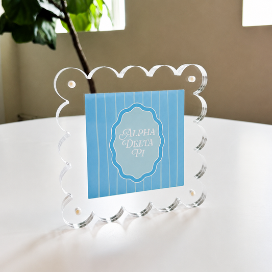 The Darling Effect’s Alpha Delta Pi Square Scalloped Acrylic Frame displays a blue “Alpha Delta Pi” card in white script, placed on a white table next to a leafy green plant and a window.